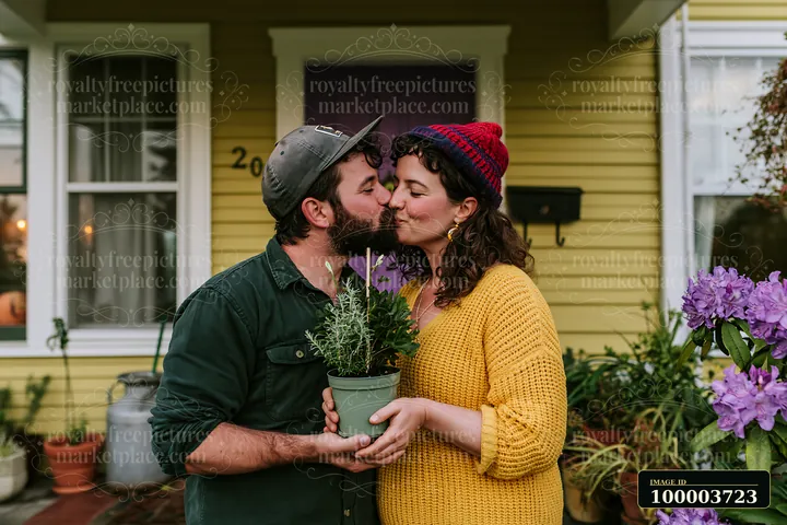Partners kiss while holding a potted plant on the porch of their yellow home.