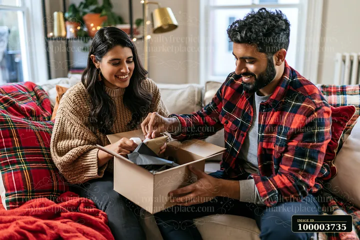 A couple is unboxing gifts while seated on a cozy sofa adorned with blankets and pillows in a festive living room filled with warm colors and decorations.