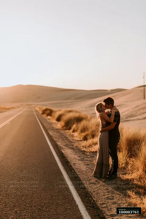 Couple kisses beside a desert road at sunset, with dunes, warm light, and expansive copy space in the sky.