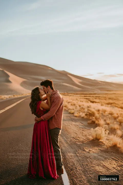 A couple is kissing on an open road surrounded by desert scenery and sand dunes during sunset.