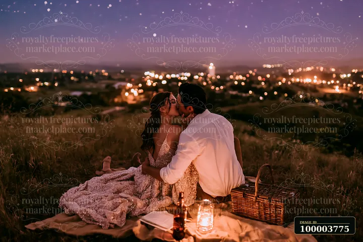 Couple kisses during a candlelit picnic in a grassy field beneath a starry evening sky.
