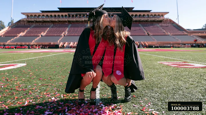 Two graduates kiss on a football field during a sunny graduation celebration, surrounded by confetti and stadium seating.