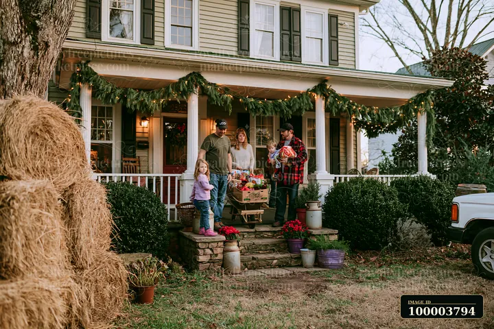 A family stands on the porch of a country house, carrying holiday decorations and gifts, with pine garlands adorning the entrance.