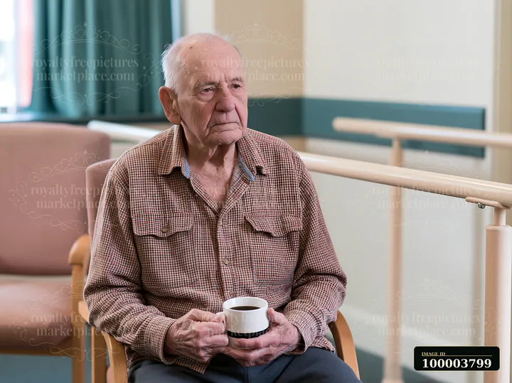 An elderly man sits in a care facility holding a cup.
