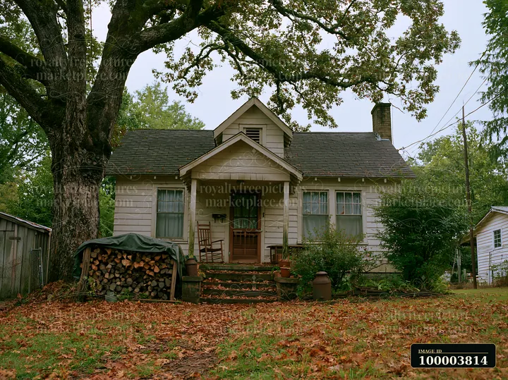 Small rural home beneath mature oak trees, with a front porch, covered woodpile, and fallen leaves in the yard.