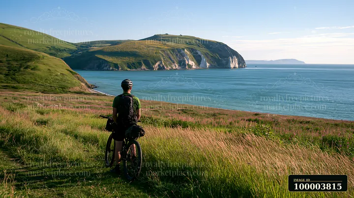 Cyclist with a mountain bike overlooks flower-covered white cliffs across calm coastal water.