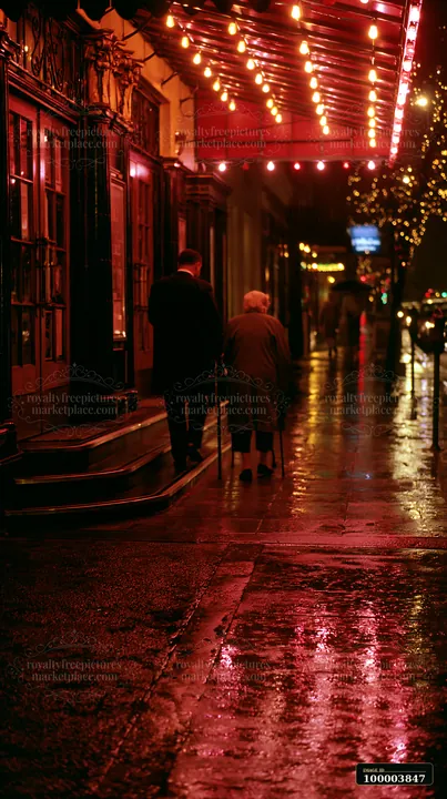 Two people walk beside a decorated storefront as Christmas lights reflect on wet pavement at night.