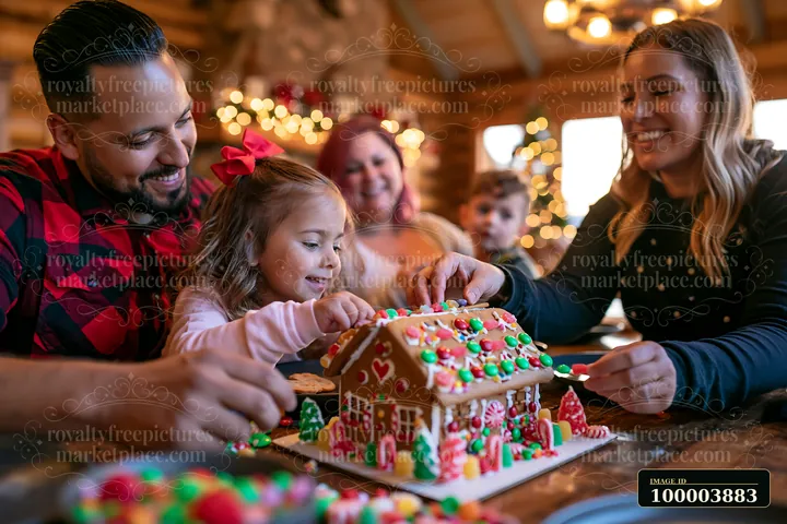 Family members decorate a colorful gingerbread house together at a warmly lit wooden table.