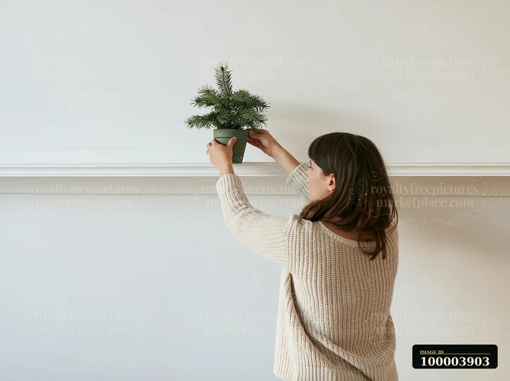 Woman places a small potted evergreen tree on a white mantel in a bright apartment.
