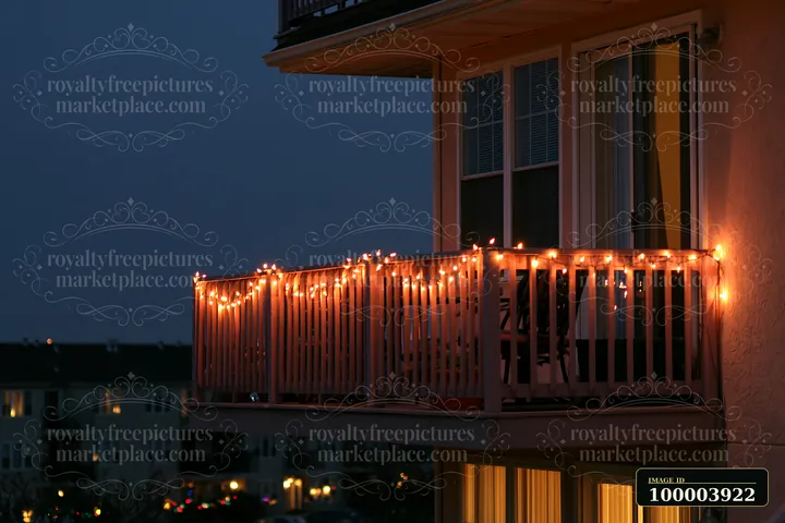 Warm Christmas lights glow along a residential balcony railing at dusk beside illuminated home windows.