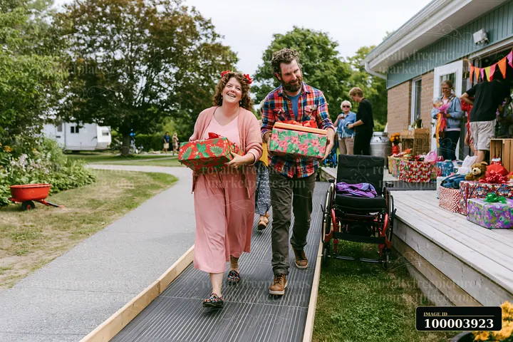 A woman and a man carry decorated gift boxes while walking on a path outside a building.