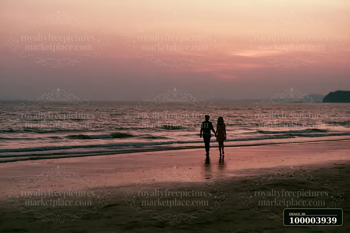 A couple walks hand in hand along the shore as the sun sets over the ocean.