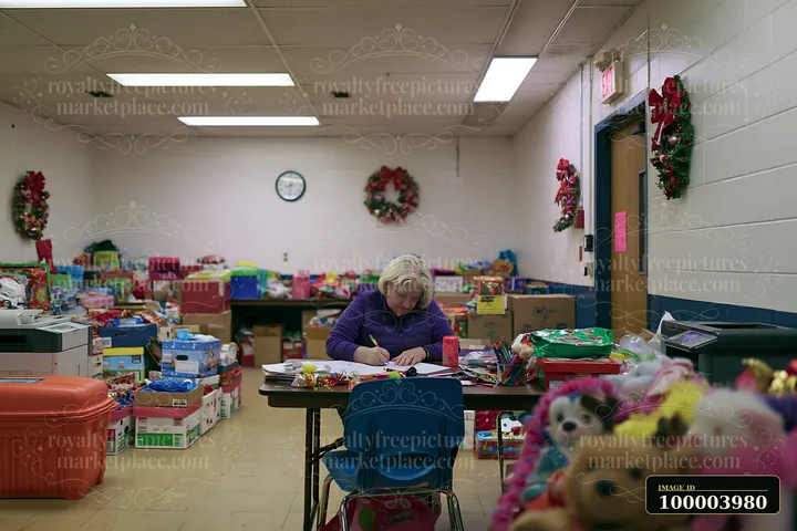 A person is writing a gift list at a desk surrounded by boxes of gifts and holiday decorations.