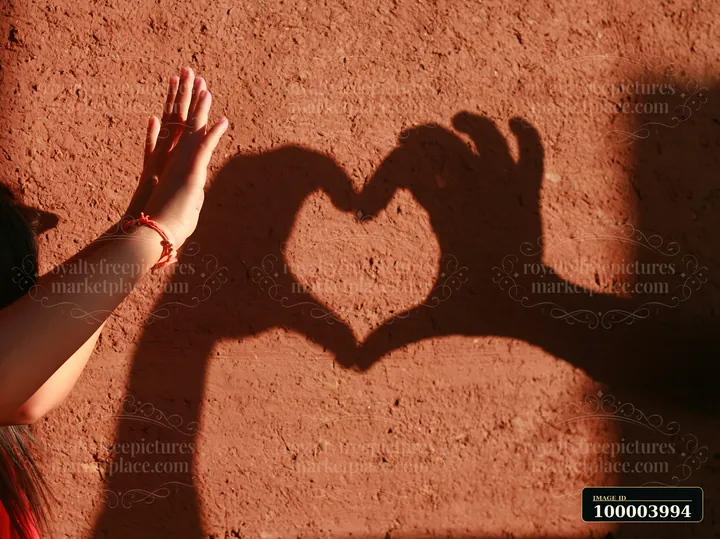A hand reaches toward a heart-shaped shadow on a sunlit textured wall.