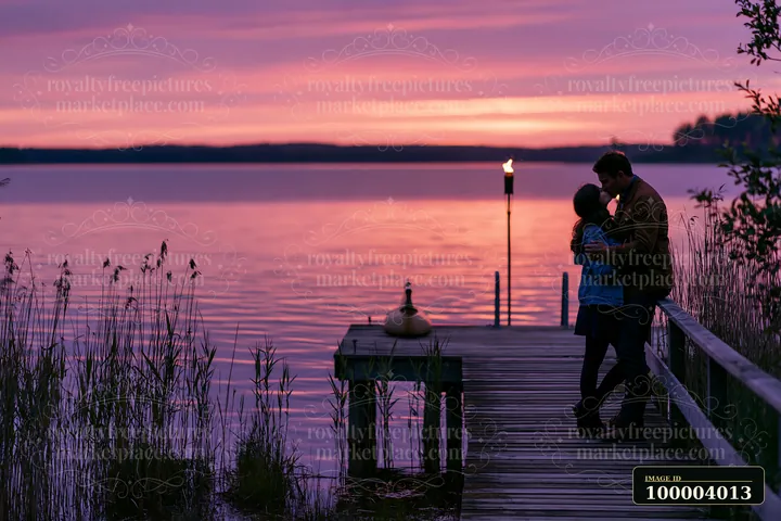 A couple kisses on a dock as the sun sets over the lake, creating a colorful sky.