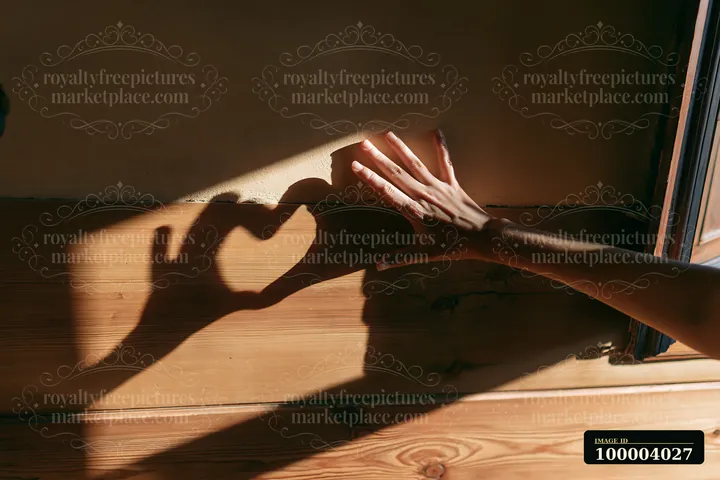 A hand near a window casts a heart-shaped shadow across a sunlit wooden wall.