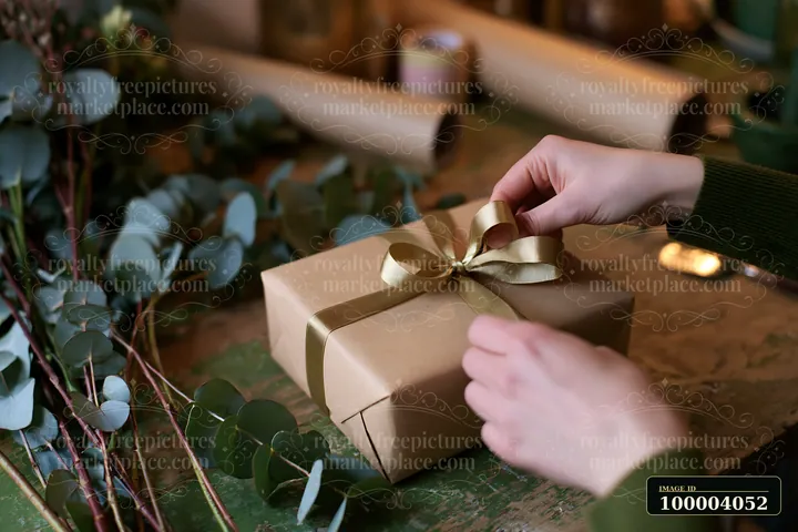 Hands tie a gold ribbon around a wrapped gift box on a rustic table with eucalyptus branches.