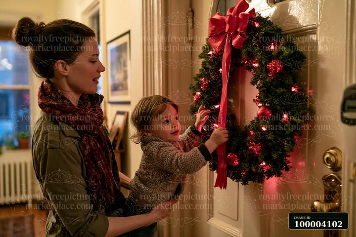 A child helps their mother hang a Christmas wreath adorned with lights and ornaments on an apartment door.