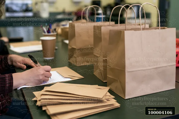 Person writes on a gift list at a desk beside paper shopping bags and envelopes.