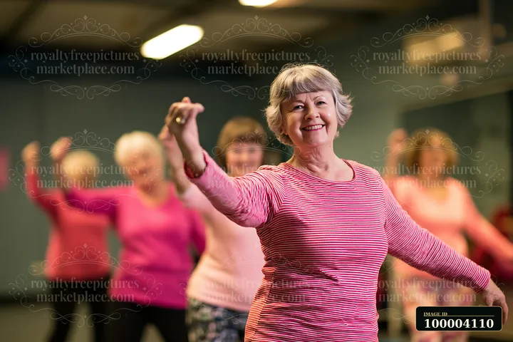 Smiling senior woman raises her arms while women practice dance fitness in a warmly lit studio.