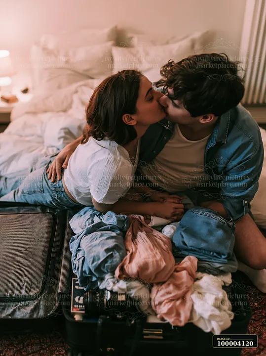 Two partners share a tender kiss while sitting together beside open luggage in a cozy bedroom.