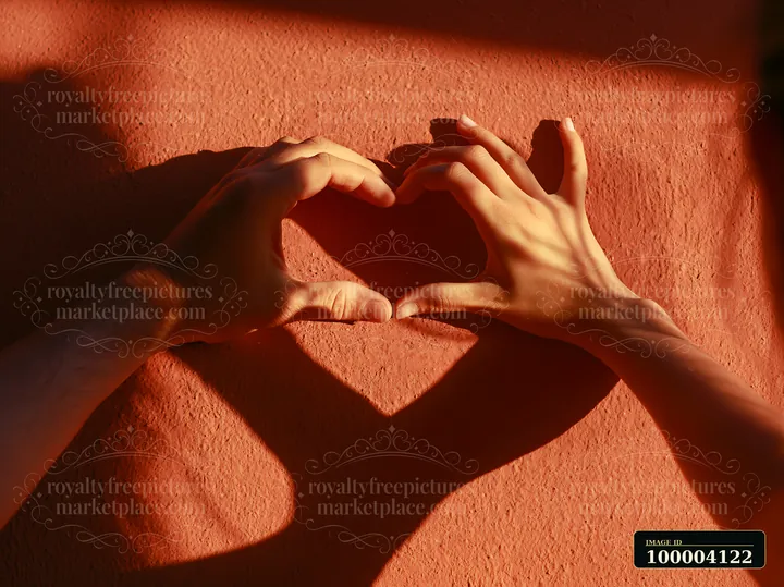 Two hands create a heart shape, casting a shadow on a textured wall in warm sunlight.