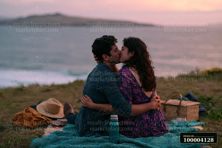 A couple sits on a blanket by the beach, sharing a kiss while surrounded by nature.