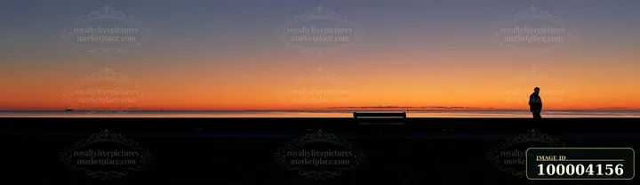 A silhouetted person stands beside a bench overlooking calm water beneath an orange and purple sunset sky.