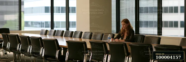 Businesswoman reviews documents alone at a conference table in a bright office meeting room.