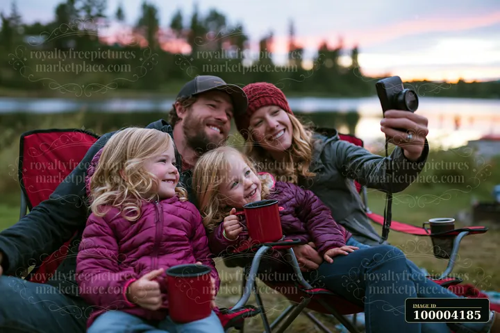 A family sits together in camping chairs by the lake, smiling as they take a selfie.