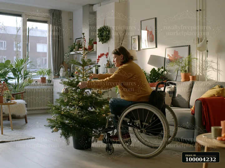 A person in a wheelchair decorates a Christmas tree in a small apartment filled with plants and holiday decorations.
