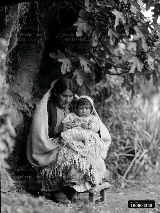 Mother and child sit wrapped in warm blankets beneath a leafy tree in a woodland setting.