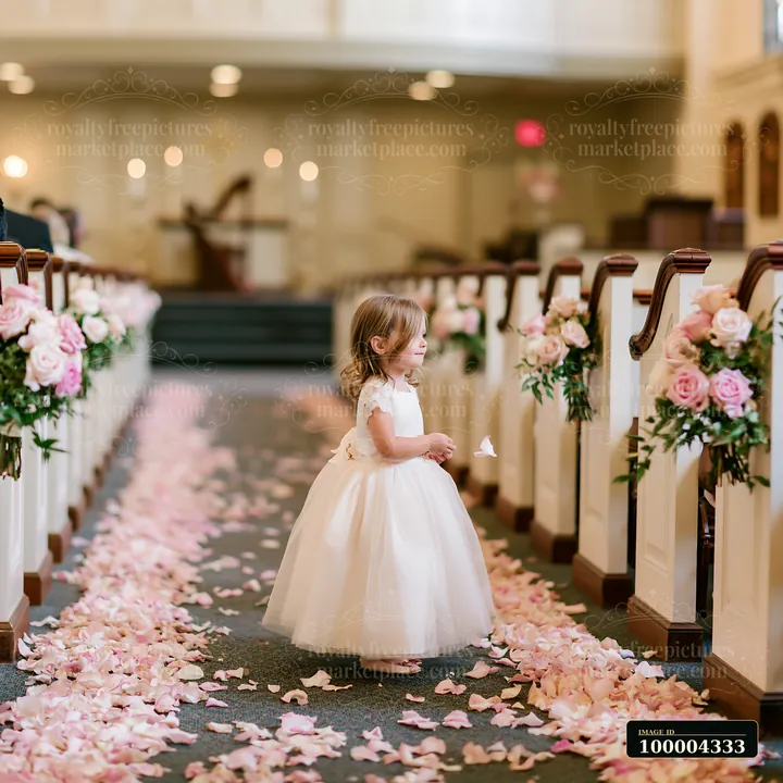 Young flower girl in a white dress walks through a church aisle covered with pink petals.
