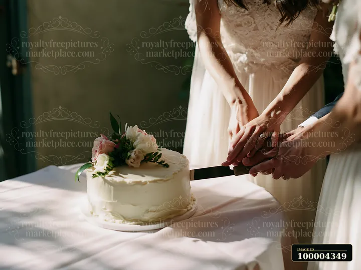 Couple cutting a flower-decorated wedding cake at a sunlit table with open copy space on the left.