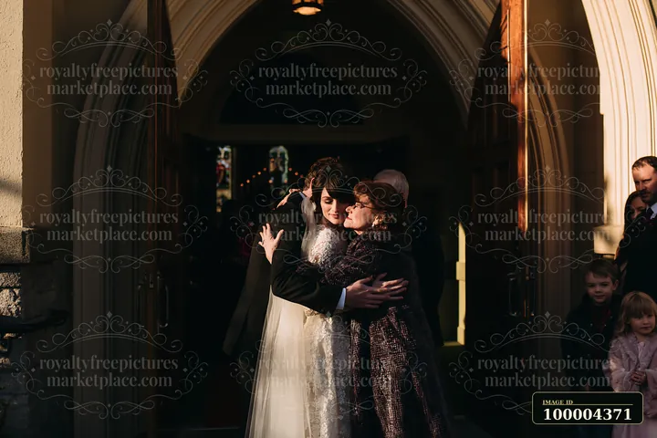 Newlywed couple embraces family beneath a sunlit church doorway arch while wedding guests gather nearby.