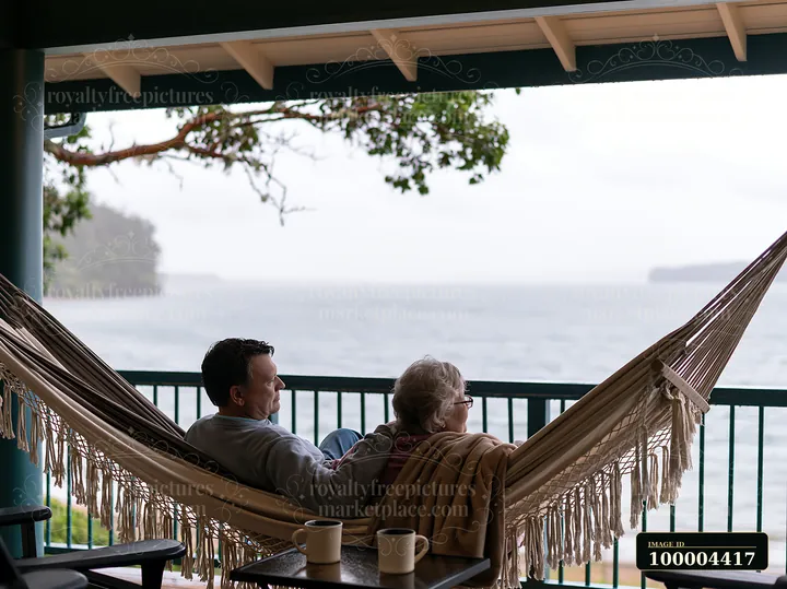 An older couple is seated together in a hammock on a porch overlooking the water.