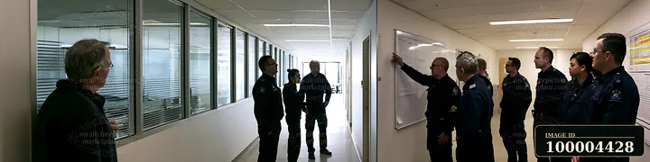 Police chief points at an operations planning board while uniformed officers observe in an office hallway.