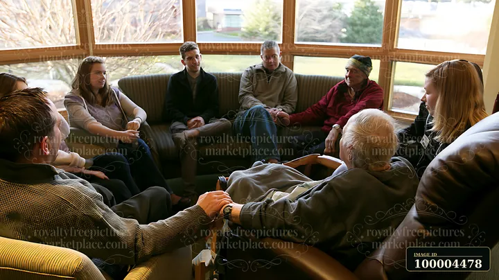A family sits together in a living room, holding hands while gathered around a hospice patient seated in a chair.