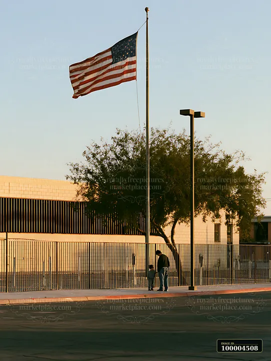 American flag waving beside a fenced school building with two people walking on the sidewalk.