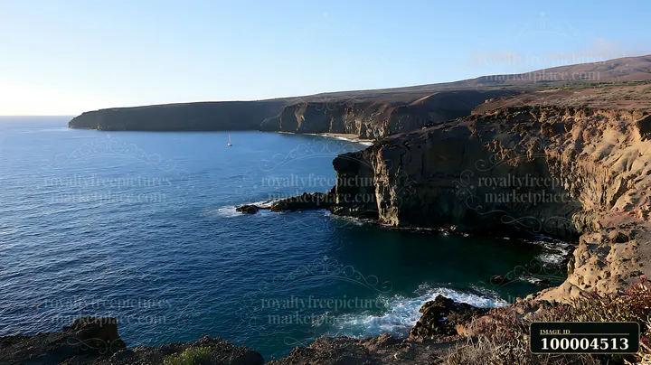 Sunlit volcanic cliffs overlook a calm blue ocean beneath a clear sky.