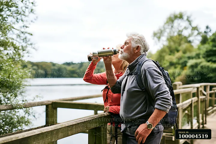 Older couple with backpacks stands on a wooden footbridge beside a lake, looking through binoculars.