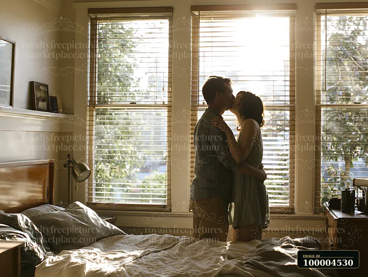 Couple kissing beside large sunlit windows in a bedroom with an unmade bed.