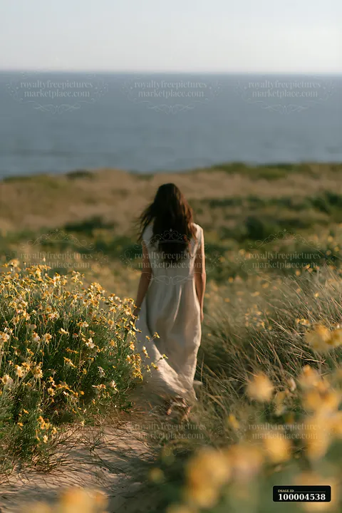 A woman walks along a sandy path lined with bright flowers, leading towards a wide ocean view under clear skies.