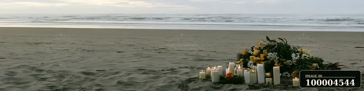 Flowers and lit candles form a memorial on a sandy beach beside the ocean.