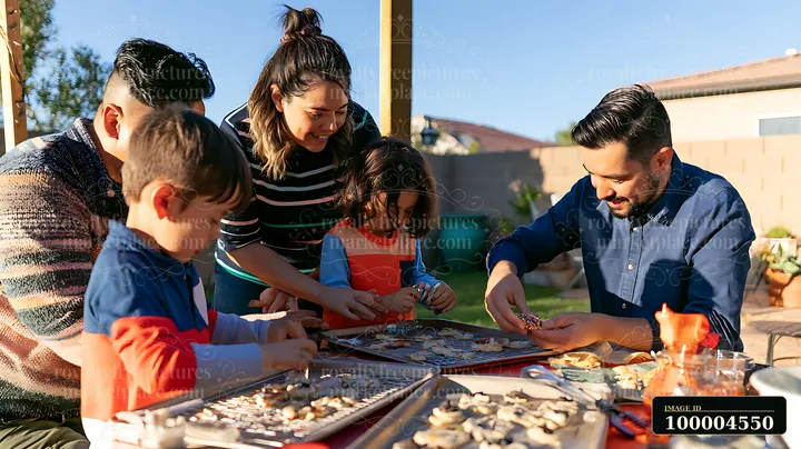 Parents and three children decorate cookies together at an outdoor table on a sunny day.