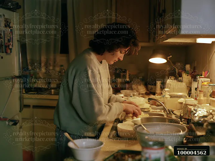 Woman prepares food at a cluttered kitchen counter under warm evening light.