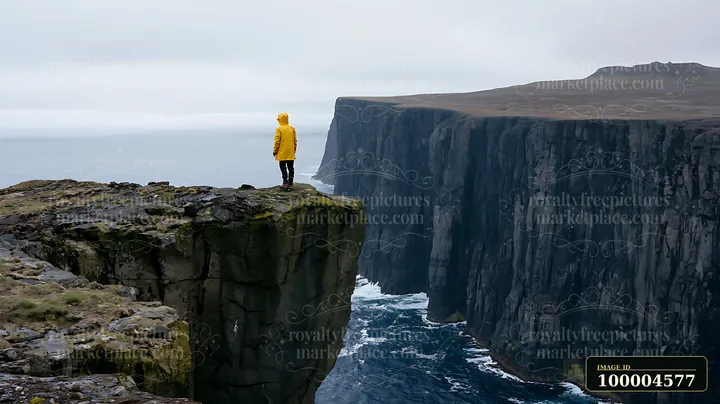 Person in a yellow raincoat stands on a rocky cliff above ocean water between towering sea cliffs.