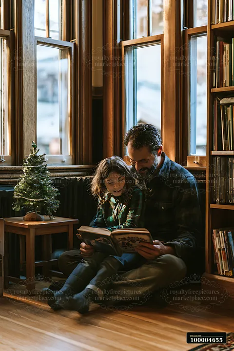 A father and daughter sit on the floor by a window to read a book together.